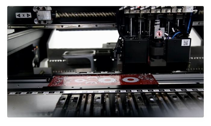 Electronics manufacturing machine with red printed circuit board positioned on assembly line track.